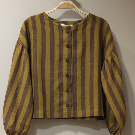 A striped long-sleeve top with a button-up front, featuring alternating mustard yellow and dark brown horizontal stripes, hanging on a beige hanger against a plain beige wall.