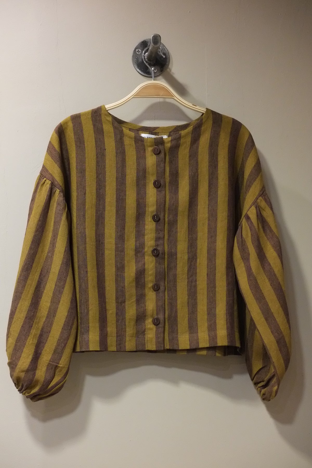 A striped long-sleeve top with a button-up front, featuring alternating mustard yellow and dark brown horizontal stripes, hanging on a beige hanger against a plain beige wall.