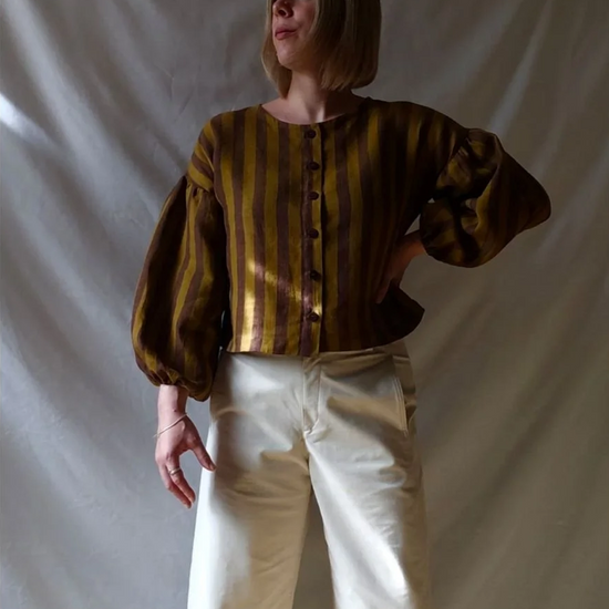 A person wearing a cropped long-sleeve blouse with vertical gold and brown stripes, paired with cream wide-leg pants, standing against a draped off-white fabric backdrop.