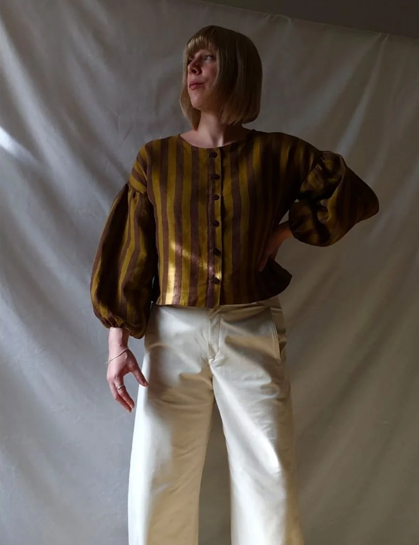 A person wearing a cropped long-sleeve blouse with vertical gold and brown stripes, paired with cream wide-leg pants, standing against a draped off-white fabric backdrop.