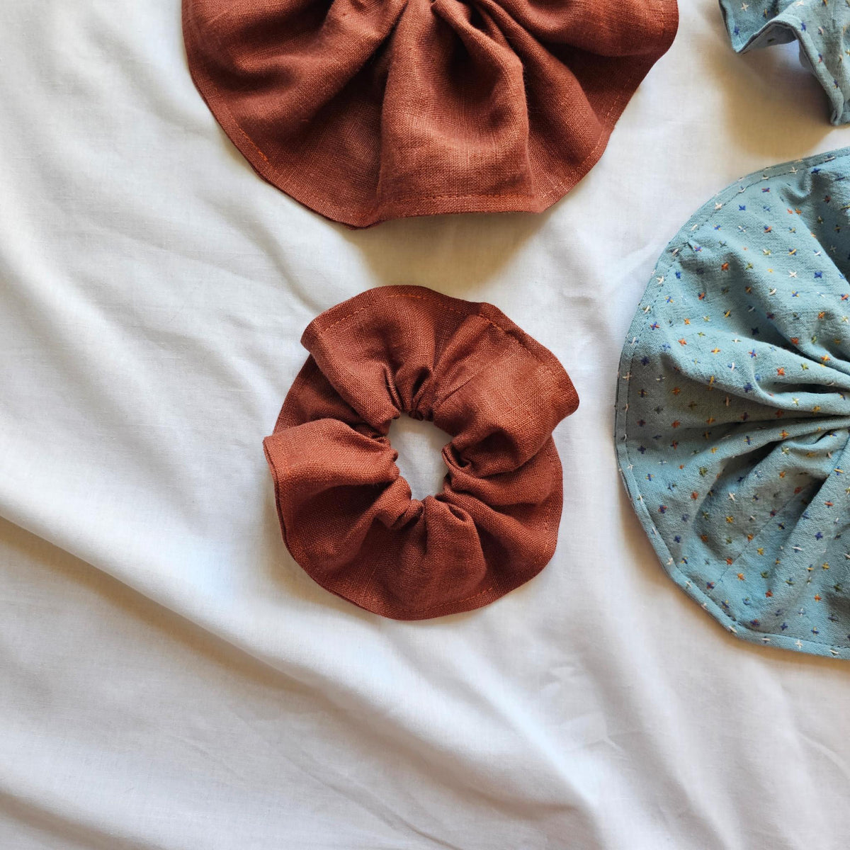 Four fabric hair scrunchies laid flat on a white fabric background: one in rust brown and one in light blue with small multicolor speckles. A partial matching blue scrunchie is visible at the top right.