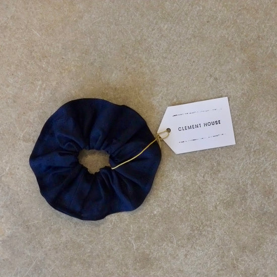 Navy blue scrunchie with a string price tag tie on a beige surface, accompanied by a 'Clement House' tag.