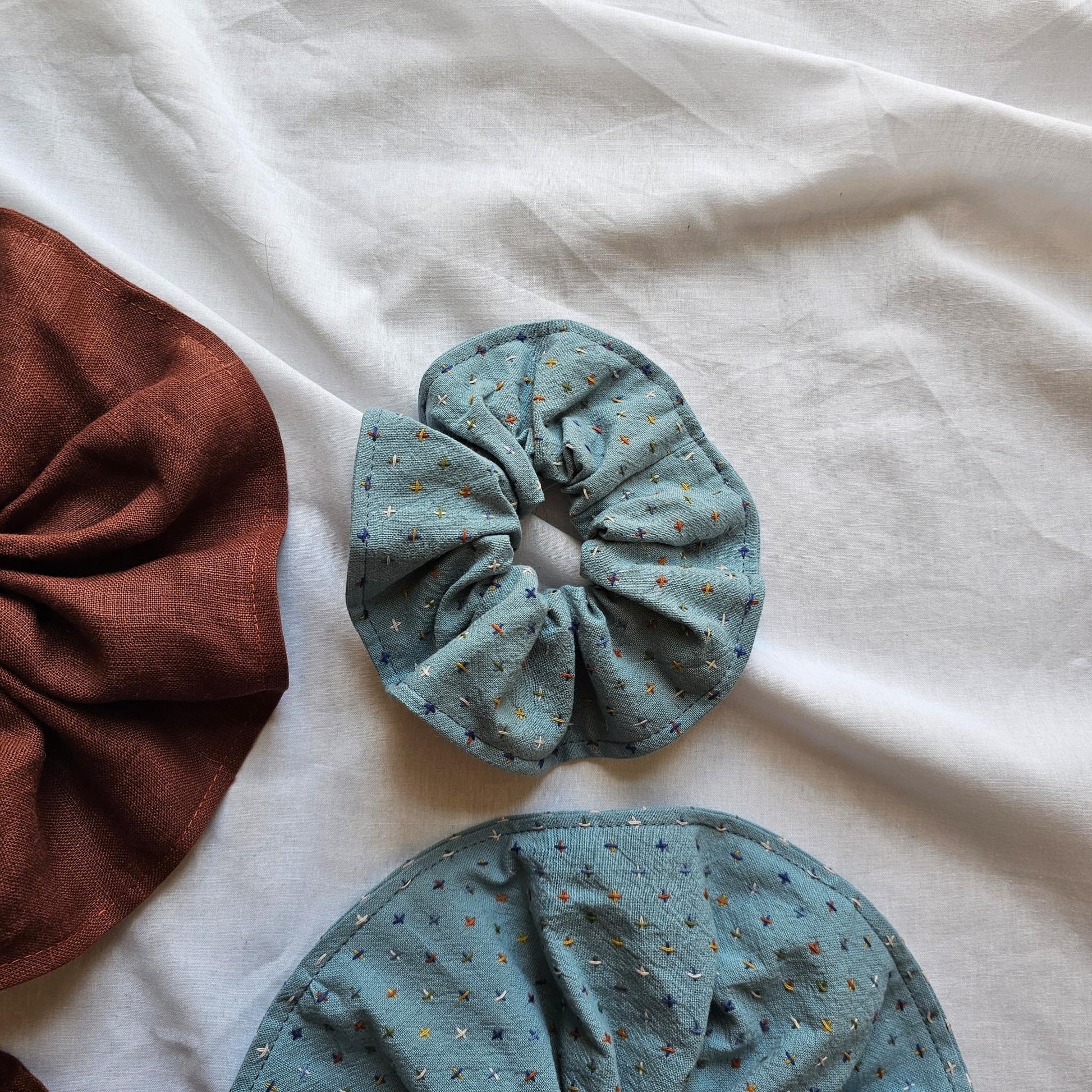 Two fabric hair scrunchies on a white fabric background, with one fully visible in a dusty blue-gray color with tiny multicolor speckles, and another partially visible on the left in a rust-brown color.