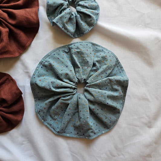 Four fabric hair scrunchies arranged on a white fabric background. The main scrunchie in the center is a dusty blue color with tiny multicolor speckles.