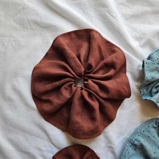 A rust-brown fabric hair scrunchie laid flat on a white sheet, with partial views of two blue patterned scrunchies at the right and bottom edges.