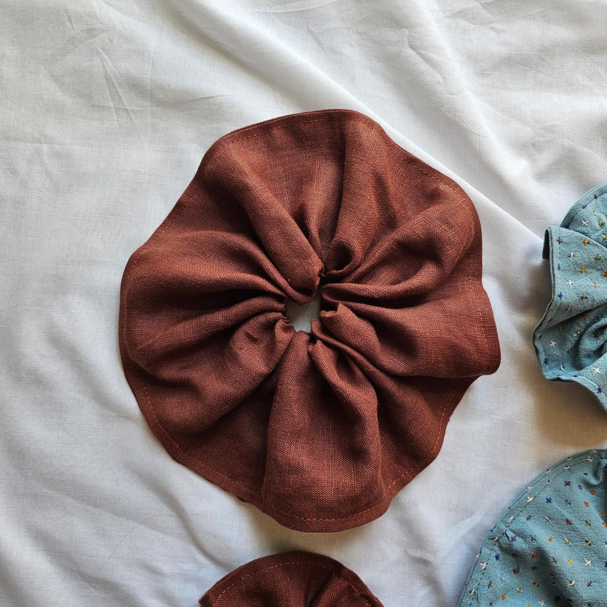 A rust-brown fabric hair scrunchie laid flat on a white sheet, with partial views of two blue patterned scrunchies at the right and bottom edges.