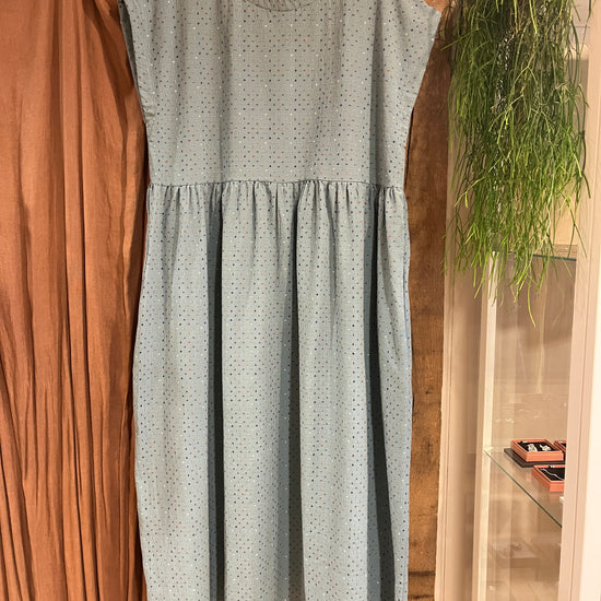 A light blue sleeveless smock dress with a small multicoloured crisscross pattern, featuring a gathered waist and tiered skirt, hanging on a wooden hanger in front of a green plant and brown curtain