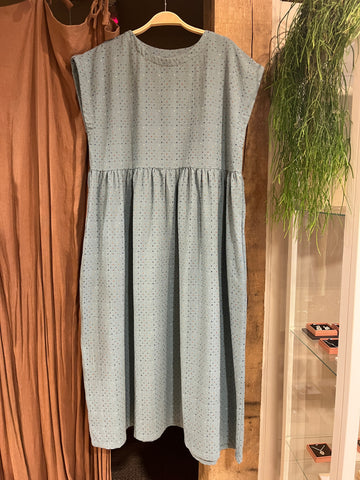 A light blue sleeveless smock dress with a small multicoloured crisscross pattern, featuring a gathered waist and tiered skirt, hanging on a wooden hanger in front of a green plant and brown curtain
