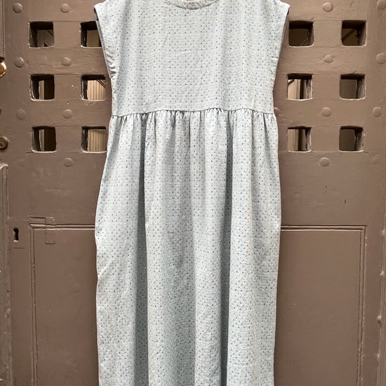 A light blue sleeveless dress with a tiny multicoloured cross stitch pattern, on a wooden hanger against a grey medieval  door