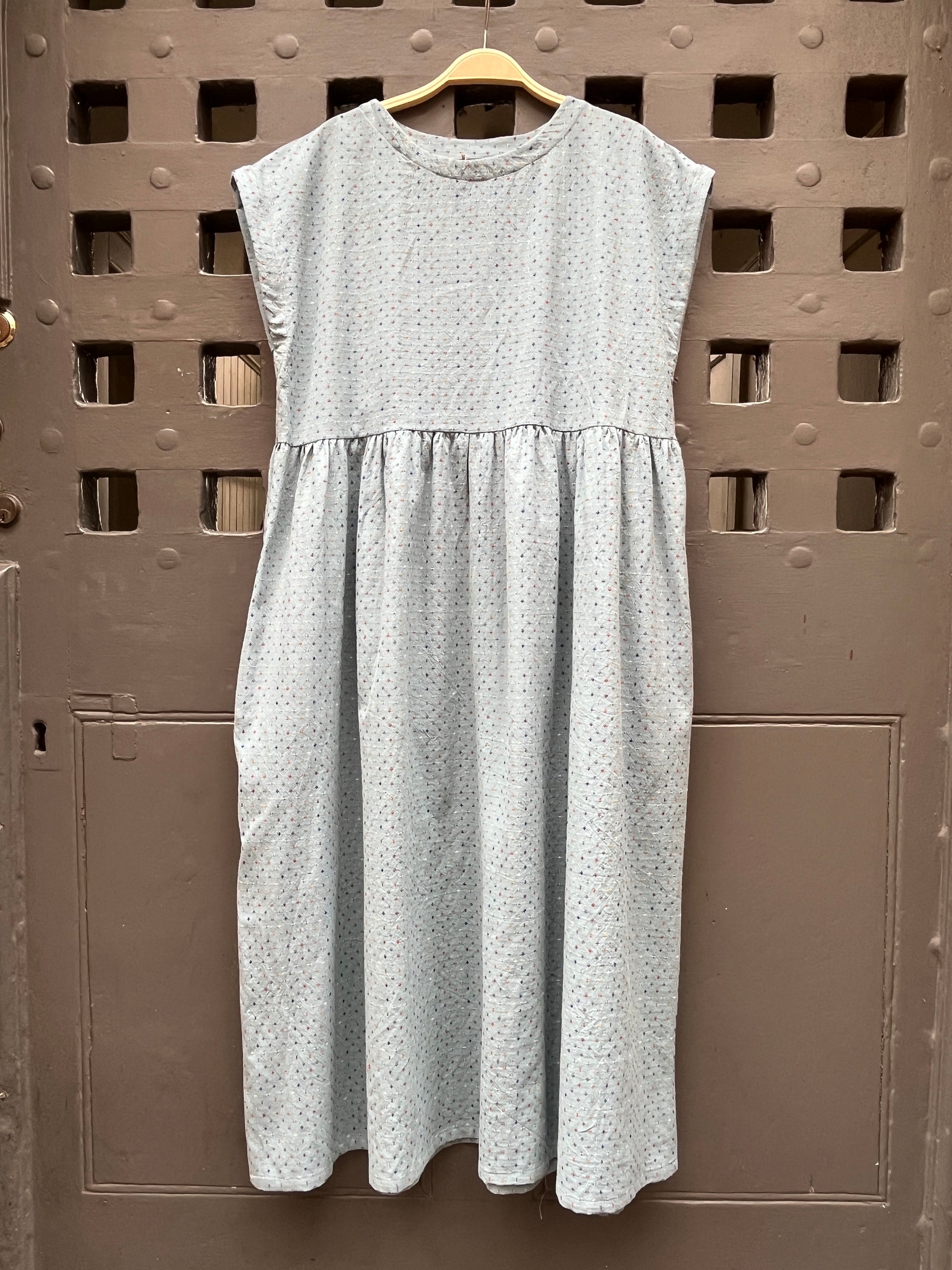 A light blue sleeveless dress with a tiny multicoloured cross stitch pattern, on a wooden hanger against a grey medieval  door