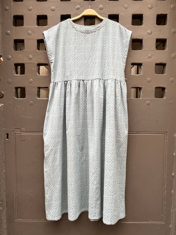 A light blue sleeveless dress with a tiny multicoloured cross stitch pattern, on a wooden hanger against a grey medieval  door