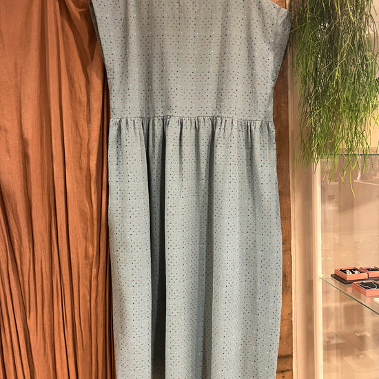 A light blue sleeveless dress with a small multicoloured crisscross pattern, featuring a gathered waist and capped sleeves, hanging against a brown curtain and wooden post