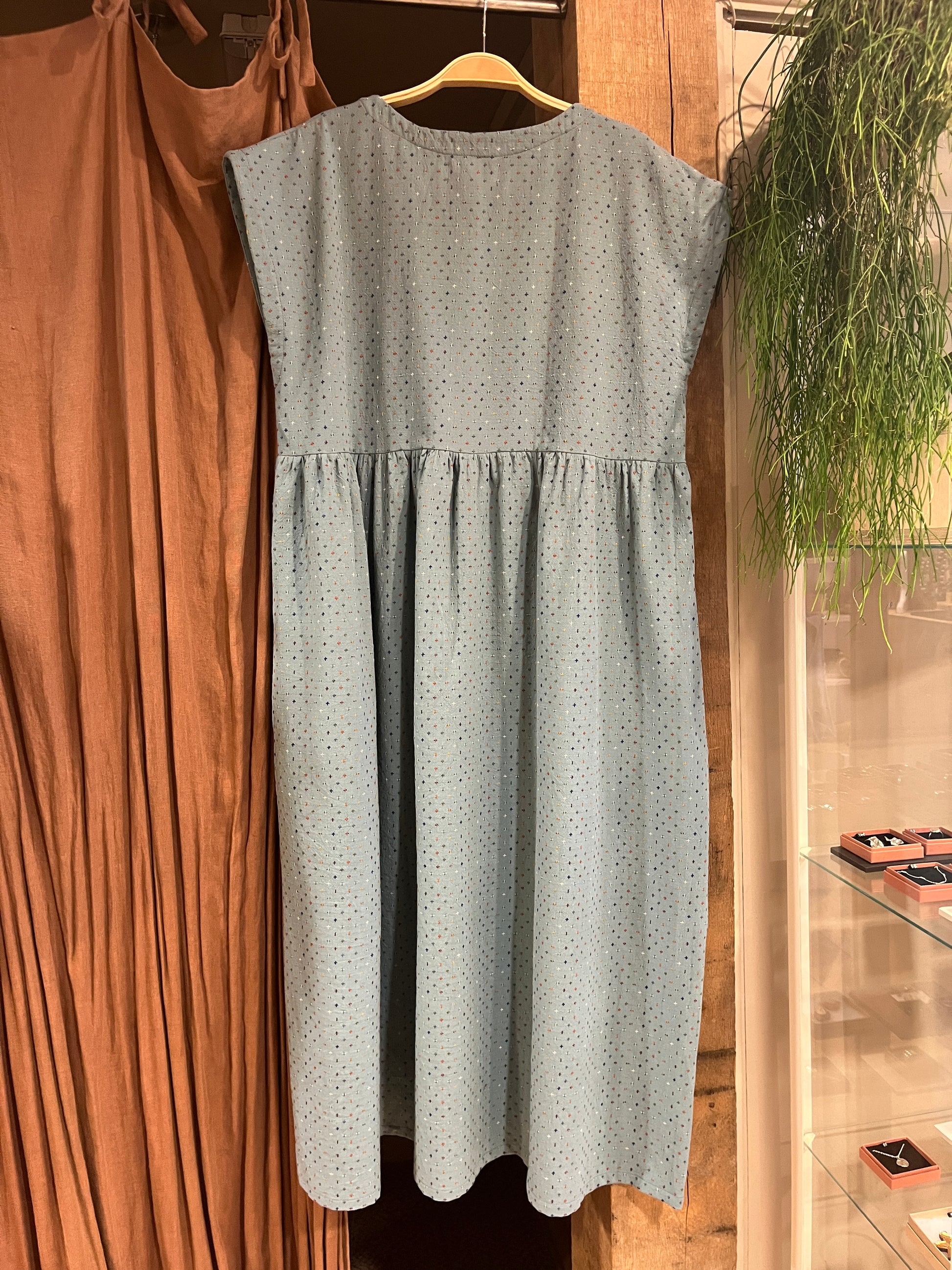A light blue sleeveless dress with a small multicoloured crisscross pattern, featuring a gathered waist and capped sleeves, hanging against a brown curtain and wooden post