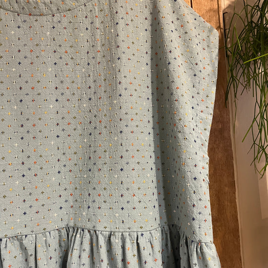 A close-up of a light blue fabric garment or textile with a small multicolour dotted pattern and ruffled detailing, hanging in front of a wooden background.