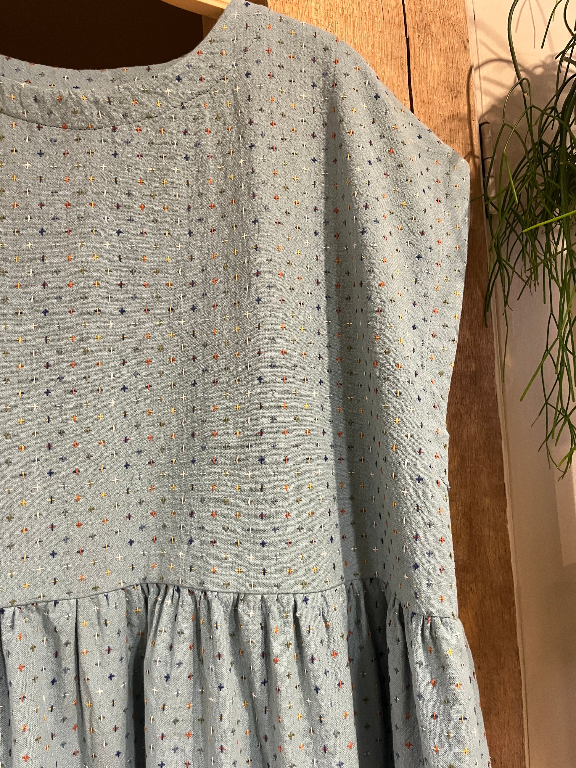 A close-up of a light blue fabric garment or textile with a small multicolour dotted pattern and ruffled detailing, hanging in front of a wooden background.