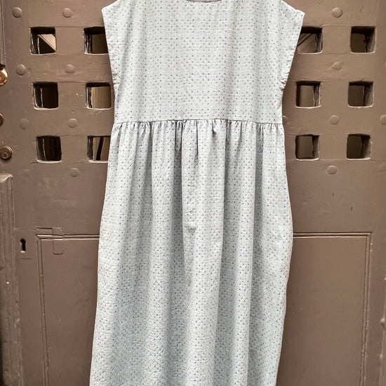 A light blue sleeveless dress with a multicoloured cross stitch  pattern, hanging on a wooden hanger against a grey rustic door.