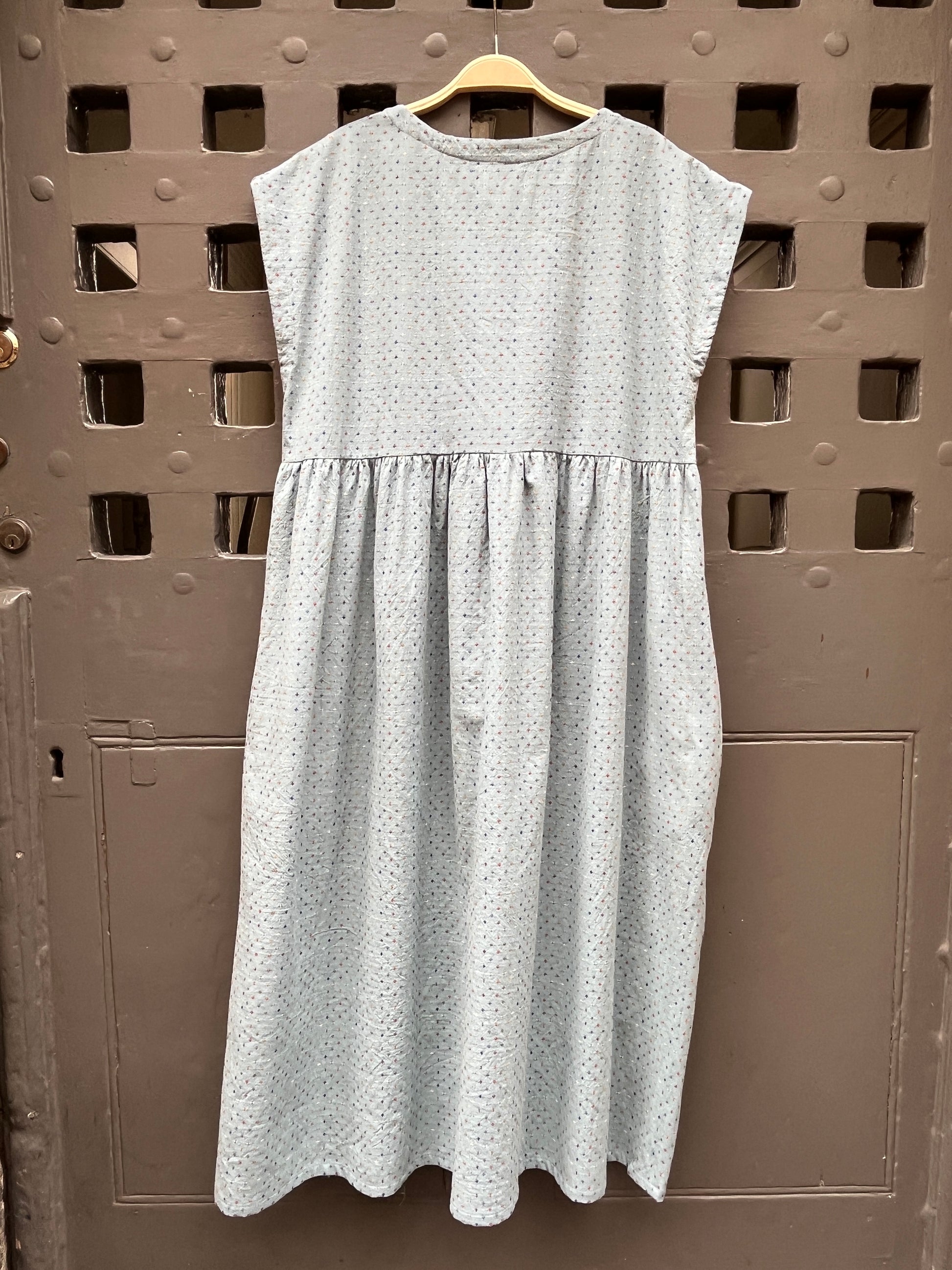 A light blue sleeveless dress with a multicoloured cross stitch  pattern, hanging on a wooden hanger against a grey rustic door.