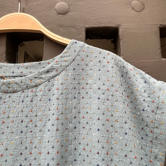 A light blue textured  top with a multicolor embroidered geometric pattern, hanging on a wooden hanger against a rustic medieval door