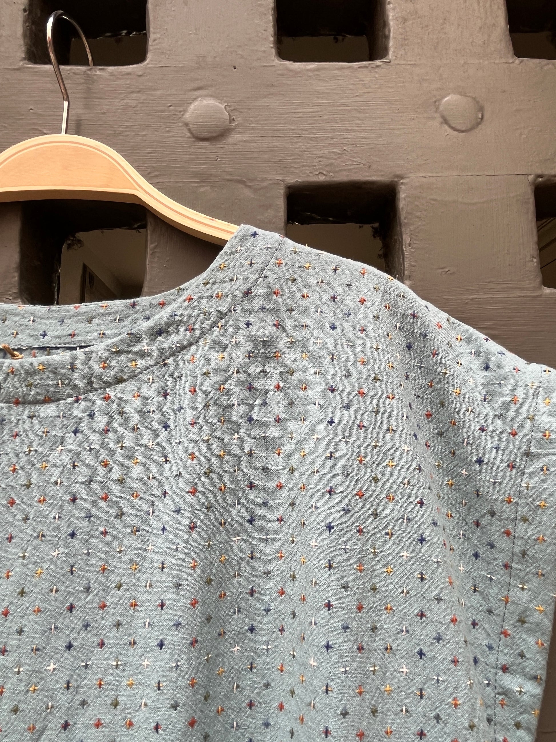 A light blue textured  top with a multicolor embroidered geometric pattern, hanging on a wooden hanger against a rustic medieval door