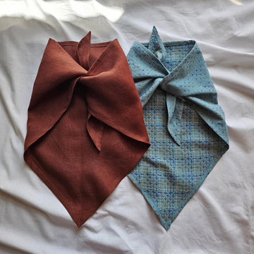 Two fabric neck gaiters or bandanas laid flat on a white fabric background. The left one is a solid rust-brown color, and the right one is a light blue patterned fabric with small dots and a subtle grid design.