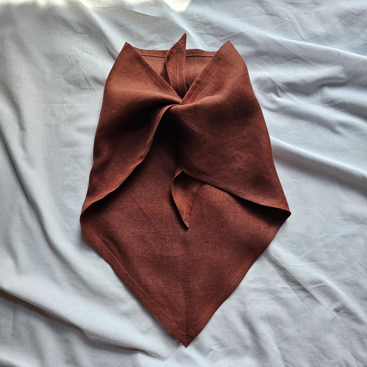 A rust-brown linen bandana or neck gaiter folded and tied in a knot, displayed on a wrinkled light gray fabric background.