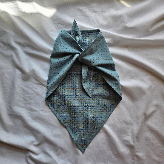 A light blue patterned neck gaiter or bandana tied in a knot, laid flat on a wrinkled white sheet.