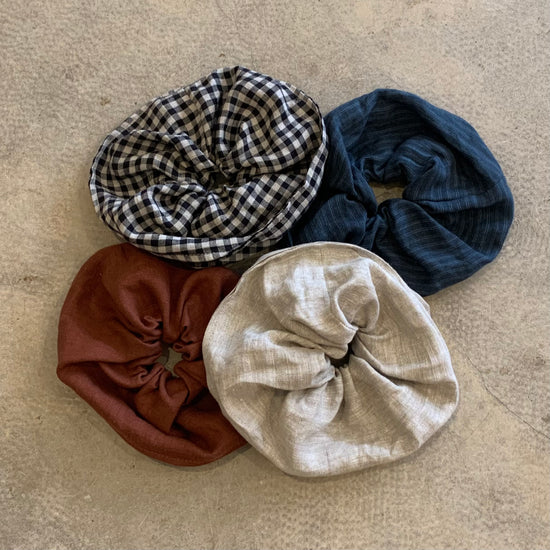 Three scrunchies displayed on a floor, one in brown, one in blue stripe pattern, one in black and white gingham and one I beige color, all made of different sustainable fabric materials.
