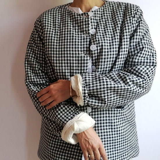 A person wearing a black and white gingham long-sleeve jacket with a high collar, white buttons, and rolled cream cuffs, standing against a plain light wall.