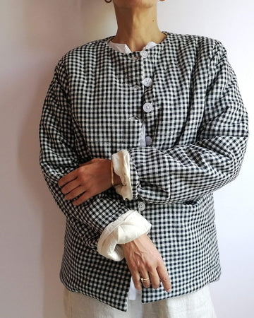 A person wearing a black and white gingham long-sleeve jacket with a high collar, white buttons, and rolled cream cuffs, standing against a plain light wall.