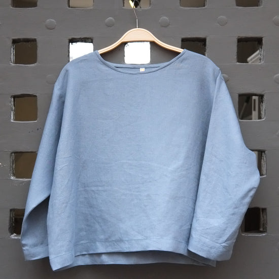 Blue round neck long sleeved linen top on a wooden hanger in front of a grey gridded door