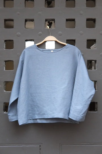 Blue round neck long sleeved linen top on a wooden hanger in front of a grey gridded door