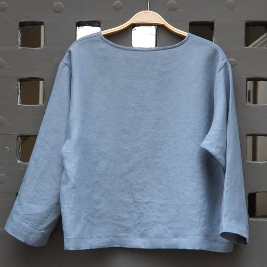 Blue round neck long sleeved linen top on a wooden hanger in front of a grey gridded door