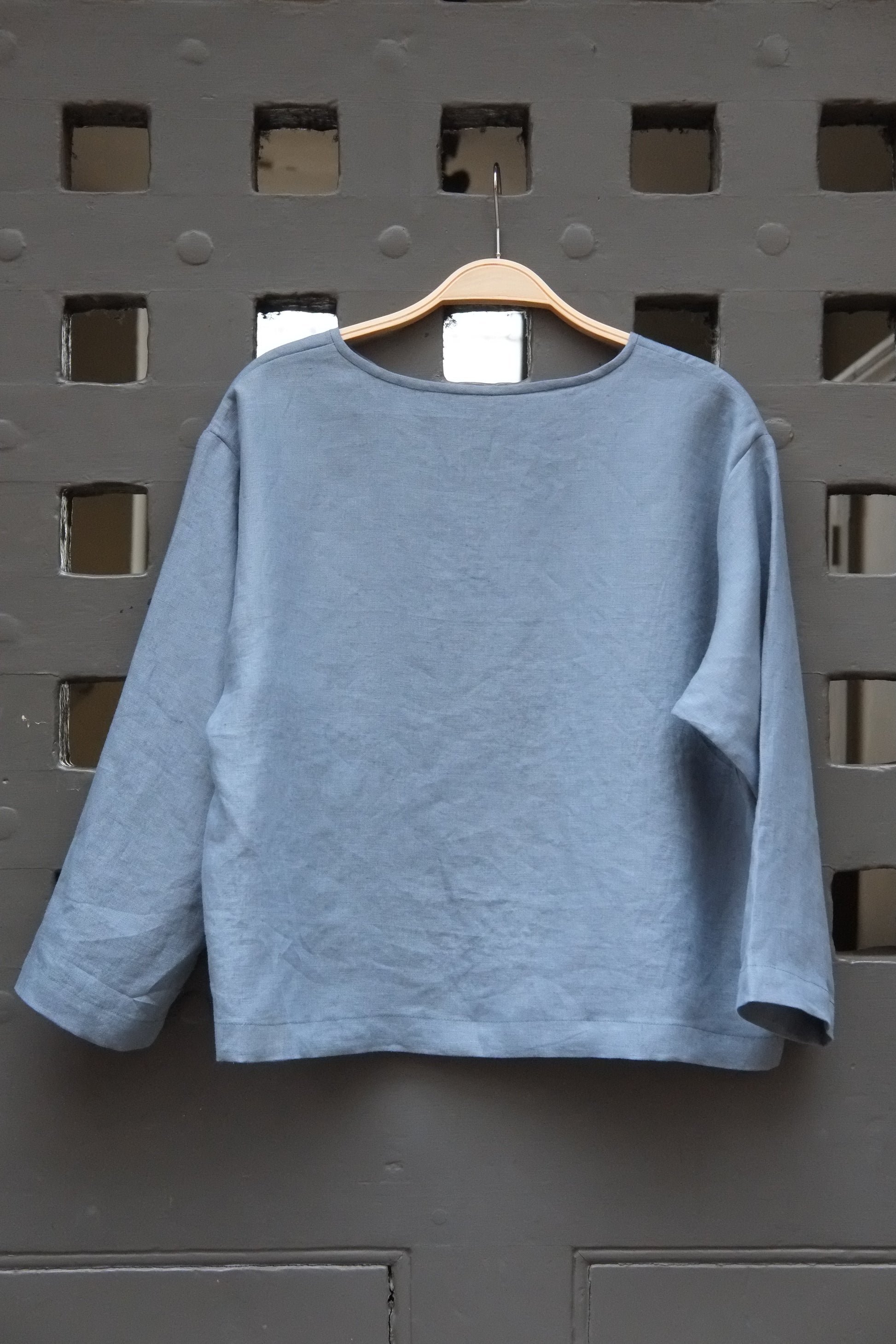 Blue round neck long sleeved linen top on a wooden hanger in front of a grey gridded door