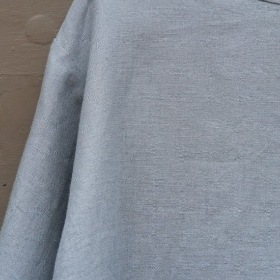 Close up shot of blue round neck long sleeved linen top on a wooden hanger in front of a grey gridded door