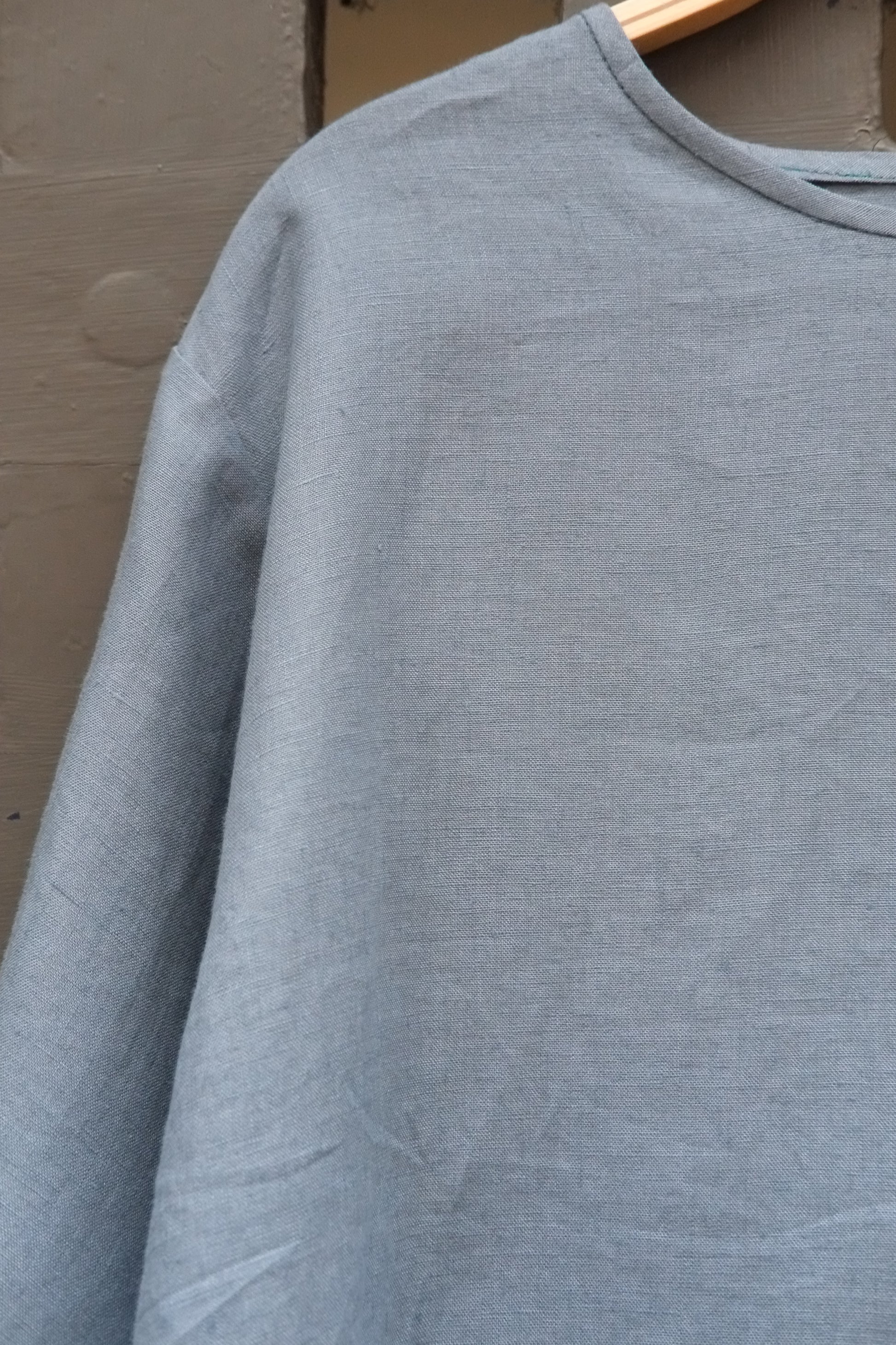 Close up shot of blue round neck long sleeved linen top on a wooden hanger in front of a grey gridded door