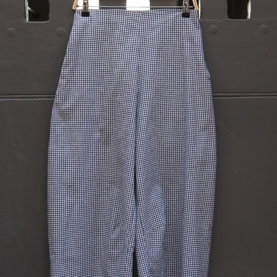 A pair of wide-leg cropped trousers in a black and white gingham pattern, hanging on a wooden hanger against a dark gray metal wall.
