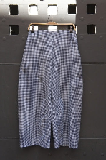 A pair of wide-leg cropped trousers in a black and white gingham pattern, hanging on a wooden hanger against a dark gray metal wall.