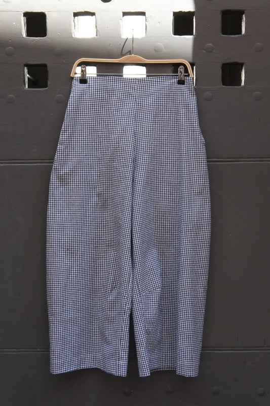 A pair of wide-leg cropped trousers in a black and white gingham pattern, hanging on a wooden hanger against a dark gray metal wall.