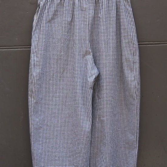 A pair of women's wide-leg cropped pants in a blue and white gingham check pattern, hanging on a wooden hanger against a dark gray paneled door.