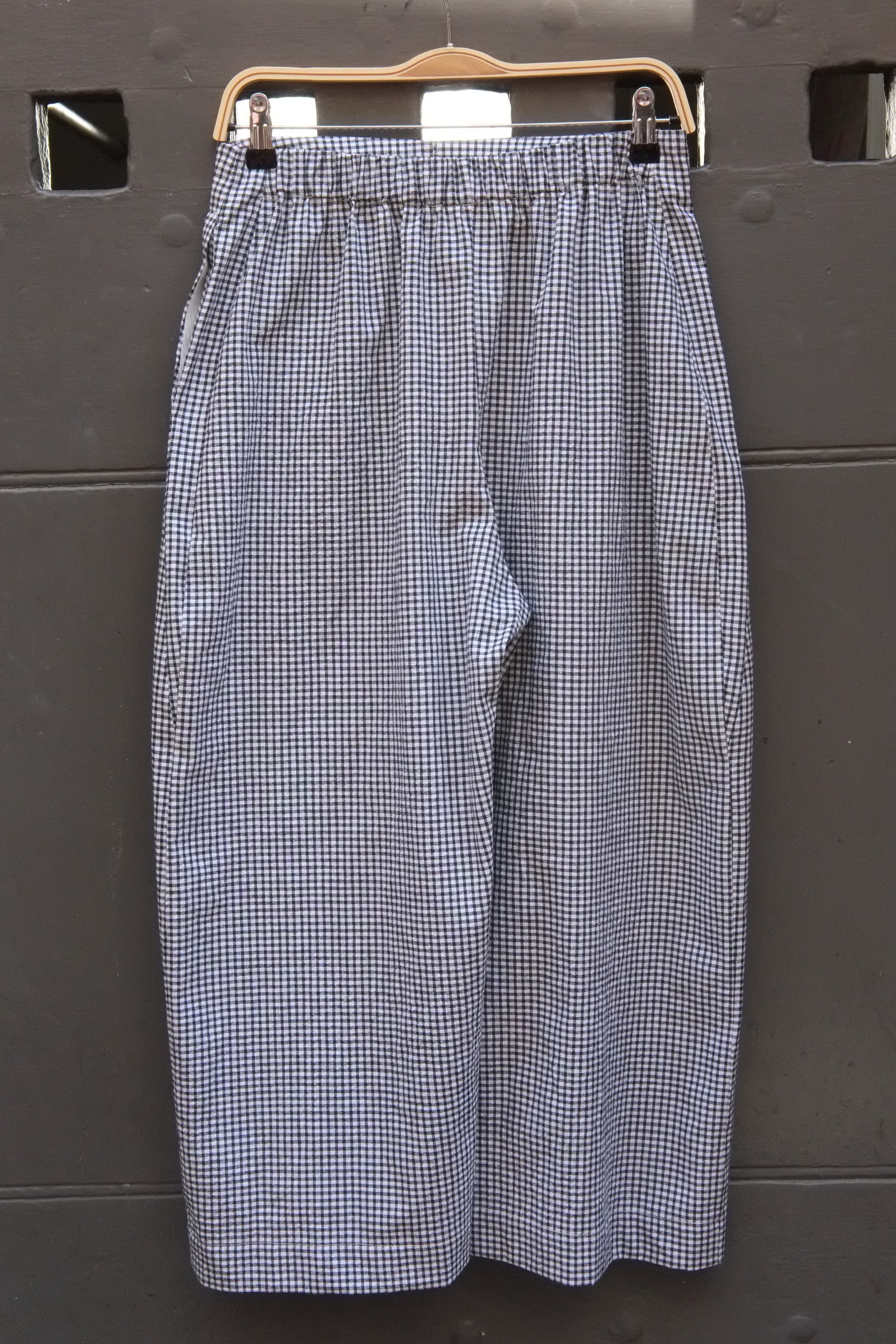 A pair of women's wide-leg cropped pants in a blue and white gingham check pattern, hanging on a wooden hanger against a dark gray paneled door.