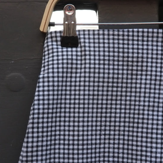 A close-up of a black and white gingham patterned skirt hanging from a wooden hanger with a metal clip against a dark wooden background.