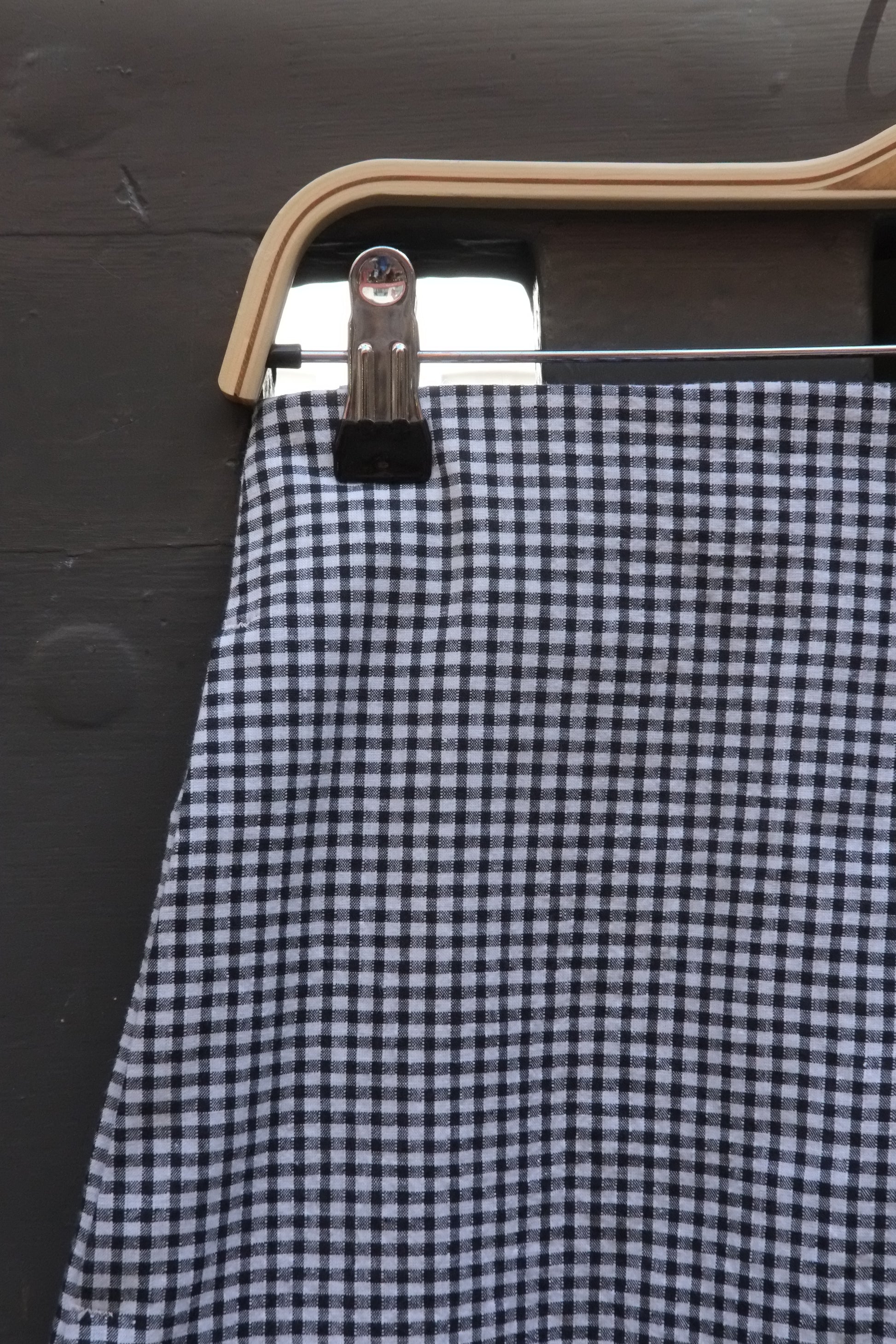A close-up of a black and white gingham patterned skirt hanging from a wooden hanger with a metal clip against a dark wooden background.