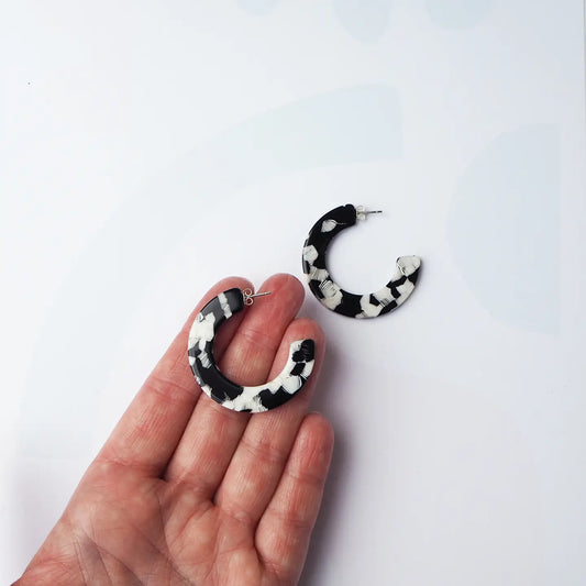 A pair of black and white acetate hoop earrings, with one earring displayed on a person's hand and the other lying on a white surface.
