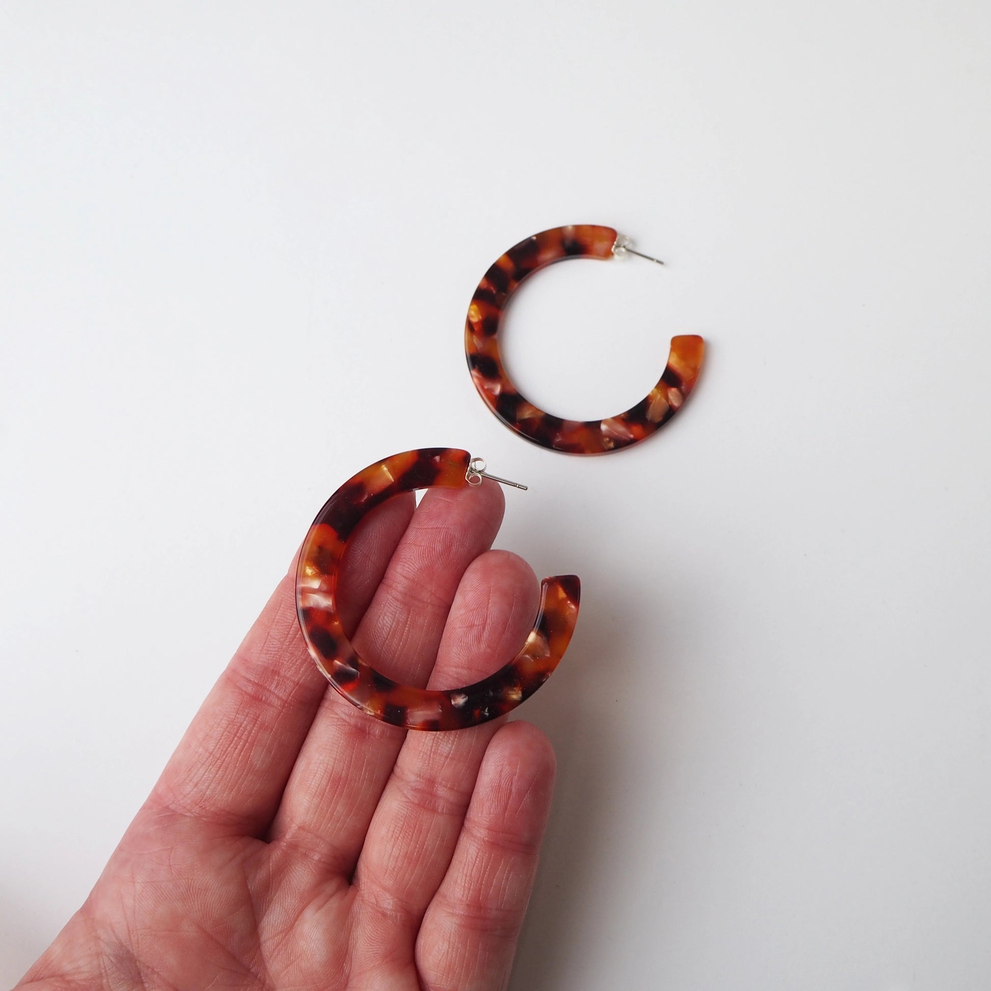 Tortoiseshell hoop earrings with a marbled brown and amber pattern, shown with one earring resting on a hand and the other floating above against a plain white background.