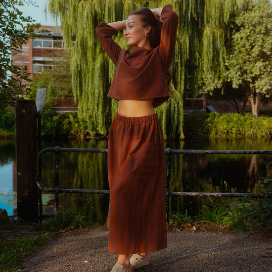 A woman stands outdoors near a canal with a large weeping willow tree behind her, wearing a rust-brown cropped long-sleeve top and matching wide-leg striped skirt. She has her hands behind her head and is wearing beige shoes.