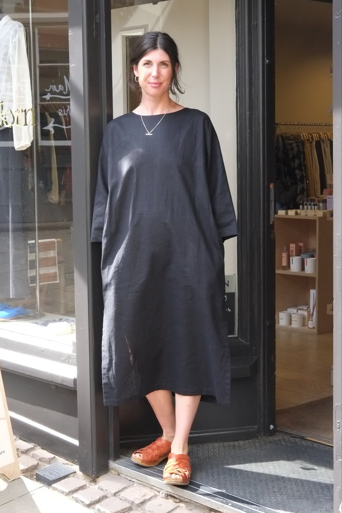 A woman wearing a loose, black midi dress stands in the doorway of a boutique. The dress has a simple round neckline and wide sleeves.
