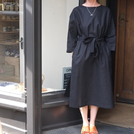 A woman wearing a black long-sleeve midi dress with a tie waist and orange shoes, standing in the doorway of a shop.