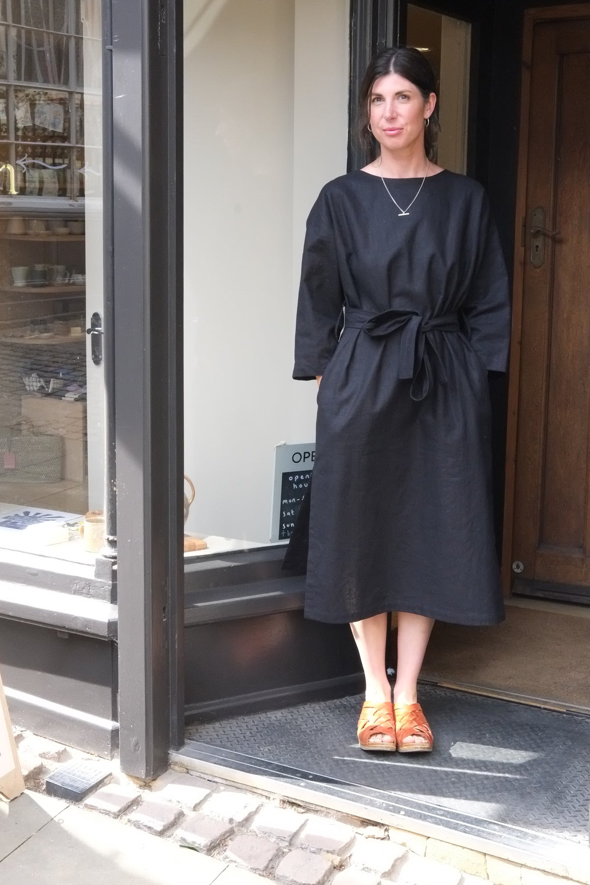 A woman wearing a black long-sleeve midi dress with a tie waist and orange shoes, standing in the doorway of a shop.