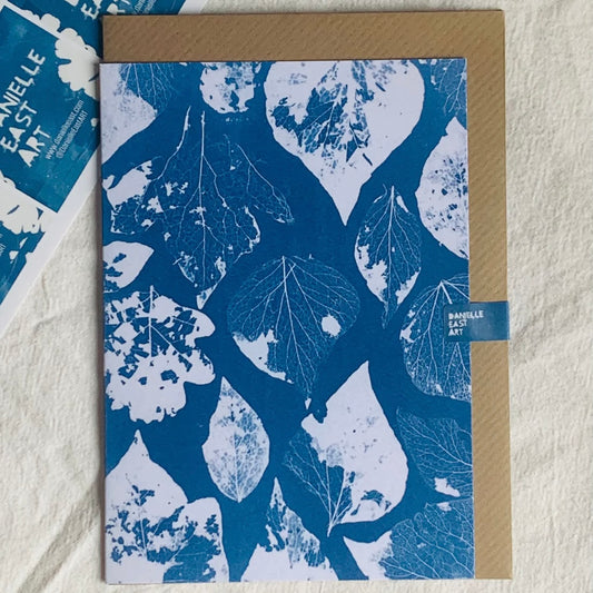 Greetings card with cyanotype blue leaf pattern and brown envelope behind on a textured surface
