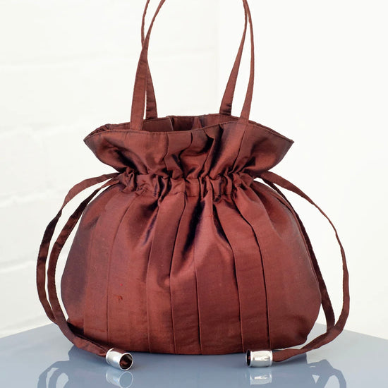 Brown silk drawstring handbag with a top handle and pleats sits on a clear surface with a white background